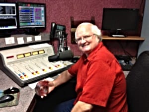 Smiling older man with white hair and glasses in a red shirt at a radio soundboard.