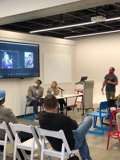 An audience watches a man speaking from a podium while a panel sits near a large screen showing two remote speakers.