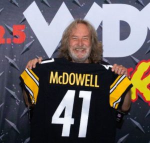 Smiling man with long hair holding up a black 'McDOWELL 41' football jersey.