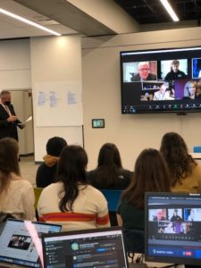 Students attend a hybrid meeting, with a speaker at left and virtual participants on a large screen at right.