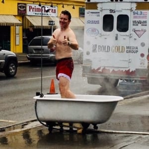 Smiling man in red shorts gives thumbs up from a bathtub being pulled in a parade.